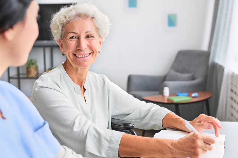 An older woman with short silver hair smiling across a table as she writes